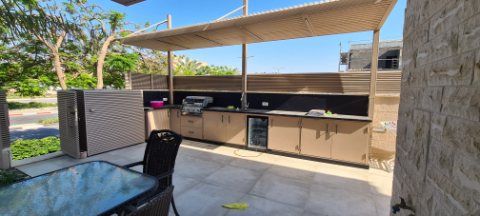 Beige aluminum pergola over an outdoor kitchen with a glass-top table in the foreground