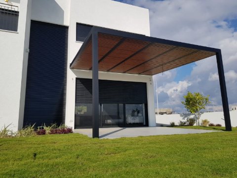 Black aluminum pergola with a wood-look roof beside a modern home, seen across the lawn