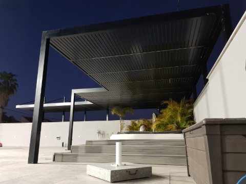Black aluminum pergola on posts over a deck and spa, seen from the side at dusk