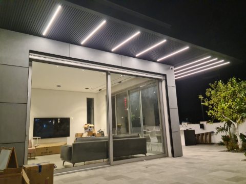 Dark louvre pergola with LED strip lights over a patio and glass sliding doors at night