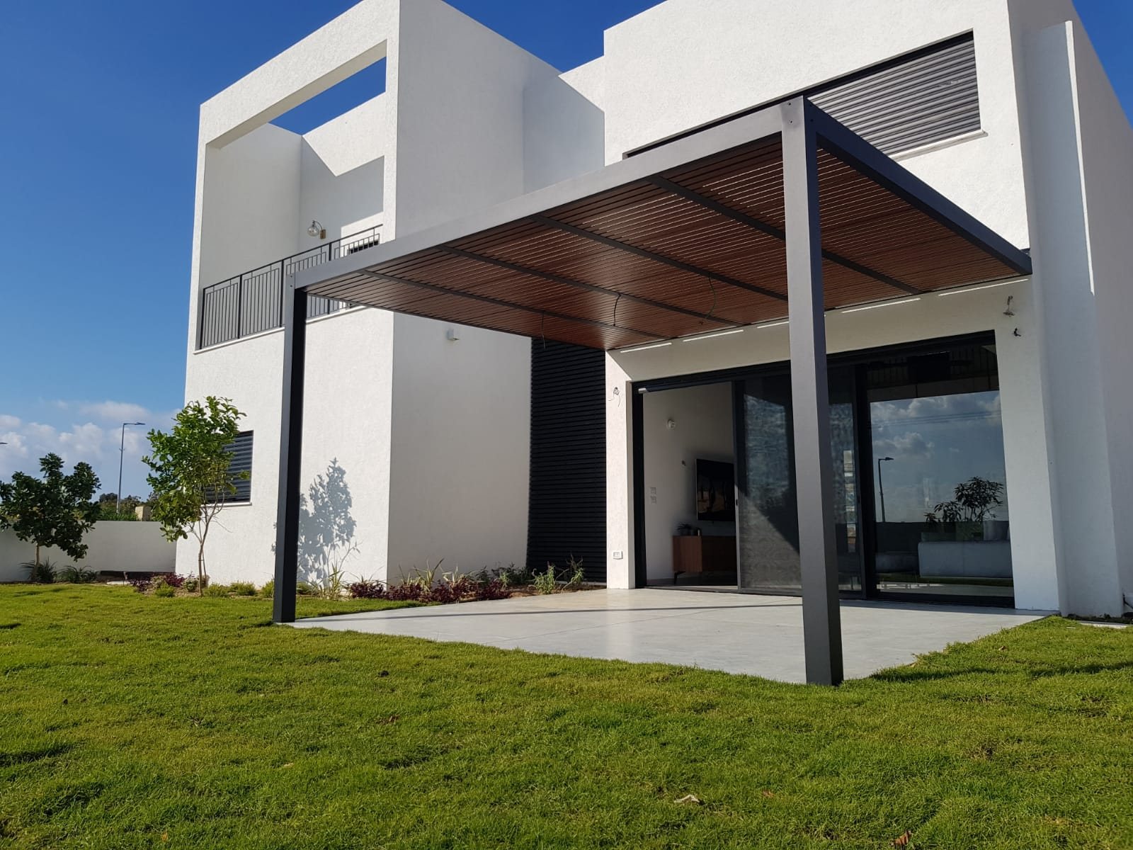 Dark aluminum pergola with a wood-look slatted roof attached to a modern white home, over a paved patio and lawn