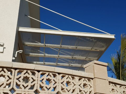 White aluminum louvre pergola fixed to a rendered wall and held by tension rods, seen from below against a blue sky
