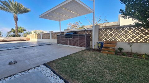 White aluminum pergola with LED lighting over an outdoor barbecue area beside a lawn and pool paving