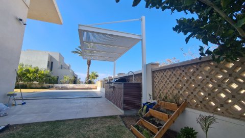 White aluminum pergola on a single post at the corner of a paved area beside a lattice wall