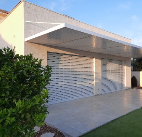 White aluminum pergola without posts, fixed to the house and supported by tension rods, over a tiled patio and artificial lawn