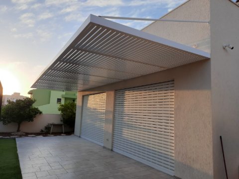 White louvre pergola without posts above a tiled patio and roller doors, photographed in late-afternoon light